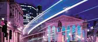 A nighttime cityscape featuring a neoclassical building with columns, illuminated by colorful lights. Light trails from passing vehicles create dynamic streaks, and modern skyscrapers are visible in the background, blending historic and contemporary architecture.