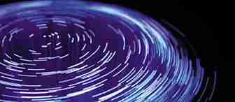 A circular, abstract light pattern with bright blue and purple streaks radiating outward on a dark background, resembling long-exposure photography of spinning lights. The streaks create a sense of motion and depth, forming a visually dynamic composition.