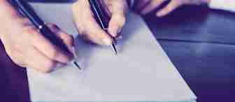 Two individuals sitting at a wooden table, with one holding a pen poised to write on a blank sheet of paper. Their hands are close together, suggesting collaboration or discussion. The focus is on their hands and the pen, with a blurred background.