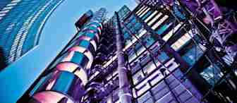 A low-angle view of modern high-rise buildings against a clear blue sky. The structures feature a mix of glass, steel, and vibrant purple lighting, showcasing architectural details with a futuristic appearance.