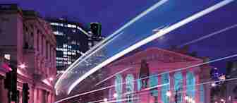 A nighttime cityscape featuring the Royal Exchange in London, illuminated with pink and blue lights. Light trails from passing vehicles create dynamic streaks in the foreground. Surrounding buildings are also visible under a deep blue and purple sky.