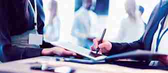 Close-up of two people engaged in a business discussion, one holding a tablet and the other using a stylus to write on it. Both individuals have nametags. A smartphone and a notebook are seen on the desk. Blurred background shows other people in conversation.