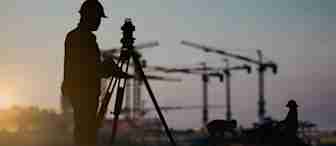 Silhouetted construction workers and surveyor with tripod equipment at a construction site during sunset. Various cranes are visible in the background, with a warm glow of the setting sun casting shadows.