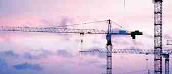 Several construction cranes are silhouetted against a pink and purple sky at dusk. The cranes' long arms intersect in a complex pattern against the backdrop of the colorful, cloud-streaked sky. The scene conveys a sense of industrial progress and early evening calm.