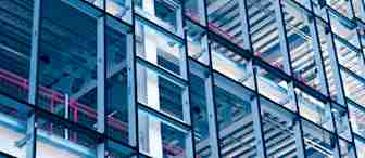 Close-up of a modern building's glass facade, revealing its structural framework. The geometric design features a grid of metal beams and glass panels, with red railings and metal conduits visible within the structure. The image has a blue tint.