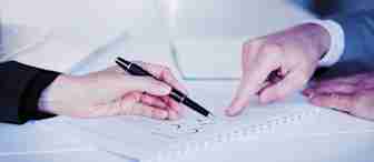 Two people, one in a dark suit and the other in a light suit, are seated at a table. One person is holding a pen and pointing to a document on the table, while the other person is pointing at the same document. The scene suggests a discussion or review of the document.