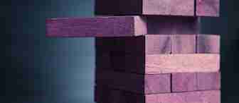 A close-up view of a tower made of purple wooden blocks in the style of the game Jenga. One block is partially pulled out from the middle of the stack, creating an imbalance. The background is slightly blurred and dark.