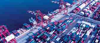 Aerial view of a busy container shipping port with multiple large red cranes, colorful shipping containers stacked in rows, and a deep blue body of water in the background. The scene shows logistics and transportation activities.