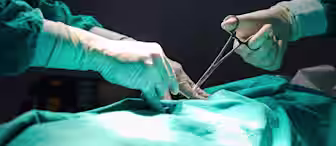Two surgeons in green surgical gowns and gloves perform a procedure on a patient, using surgical instruments under focused lighting.