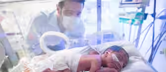 A newborn baby lies in an incubator in a neonatal intensive care unit (NICU), wearing a diaper and with medical tubes attached. An adult wearing a mask looks into the incubator, watching over the baby. Medical equipment surrounds the incubator in the background.