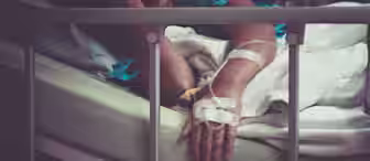 A person lies in a hospital bed with an IV drip attached to their arm. The person looks weary, with one hand resting on the bed railing and the other covering their face. The surroundings are dimly lit, emphasizing a somber mood.