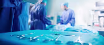 A surgeon in blue scrubs and gloves holds a surgical tool, possibly a pair of forceps, while standing next to an operating table. In the background, two other surgeons in blue scrubs and masks work on a patient under bright operating room lights. Various surgical instruments are laid out on a table in the foreground.
