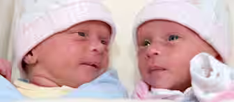 Two newborn babies lying next to each other, both wearing white hats with floral designs and pink checkered bands. The babies are wrapped in blankets and appear to be looking at each other with relaxed expressions.
