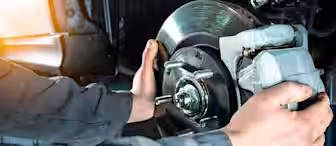 A mechanic wearing a gray jumpsuit works on a car's brake system, holding a metal brake disc and caliper. The image is well-lit, with a sun flare effect in the top left corner.