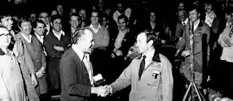 A black-and-white photo of a group of men in a workshop. Two men in the center are shaking hands, surrounded by others who are smiling and watching. The setting appears casual, with some men in overalls and work clothes.