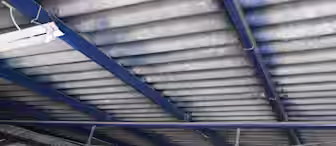 A factory ceiling with exposed blue metal beams and corrugated roofing. Several fluorescent light fixtures are mounted on the beams, illuminating the area below. There are various industrial equipment and objects partially visible at the bottom of the image.