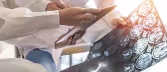 Two medical professionals in lab coats discuss and review brain scan images, with one holding a clipboard and the other holding the scans. A stethoscope and documents are visible on the table.