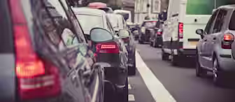 A congested city street with multiple cars lined up in traffic. The vehicles are closely packed, with buildings visible in the background. The scene captures the hustle and bustle of urban life.