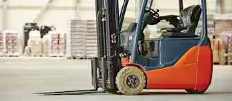 A red and black forklift is parked indoors on a concrete floor. In the background, there are stacks of boxes and pallets against a large industrial wall. The warehouse setting is well-lit.