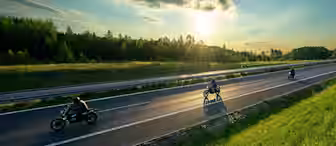Three motorcyclists ride on a smooth highway with two lanes going in each direction. The sun is setting, casting a golden glow on the scene. On either side of the highway are lush green grassy areas and dense trees in the distance under a partly cloudy sky.