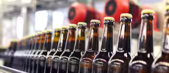 Rows of glass beer bottles with golden caps are lined up and moving along a conveyor belt in a brewery or bottling plant. The background features industrial equipment and machinery, including large red motors.