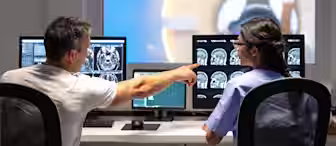 A doctor and a technician are sitting in front of multiple computer screens displaying MRI scans in a control room. The doctor is pointing at one of the screens while speaking to the technician. In the background, a patient is positioned in an MRI machine.