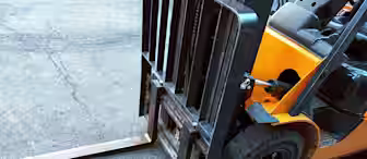 Close-up view of a forklift's front forks and wheels on a textured asphalt surface. The forklift is primarily yellow and black, with visible wear on the metal forks, indicating frequent use. The background is an urban street setting.