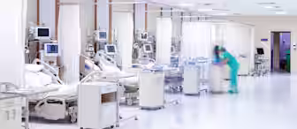 Hospital room with several patient beds, medical equipment, and monitors. A healthcare worker in green scrubs moves quickly in the foreground, while patients rest in their beds, separated by white curtains.