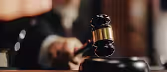 A judge in a courtroom holds a wooden gavel, poised to strike it on the sound block, with the judge’s face blurred in the background.