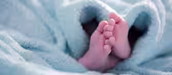 Close-up of a baby's feet peeking out from under a soft, light blue blanket. The blanket appears fluffy and cozy, enveloping the baby in warmth. The baby's tiny toes are slightly splayed, highlighting their delicate nature against the plush fabric.