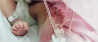 Close-up image of a newborn baby's feet in a neonatal intensive care unit (NICU). The baby has medical tubing and a white hospital bracelet on one ankle, suggesting they are receiving medical care. The baby is partially covered with a pink blanket.
