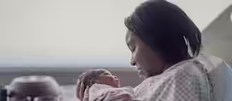 A mother in a hospital gown gently cradles her newborn baby in a hospital bed. Both mother and baby are looking at each other tenderly. The baby is wrapped in a blanket. There are hospital items in the background on a tray.