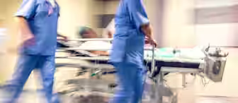 Blurred motion image of medical staff in blue uniforms pushing a patient on a hospital bed down a hallway, indicating urgency. The patient is laying down, and the surroundings are typical of a medical facility, reflecting a fast-paced emergency scenario.