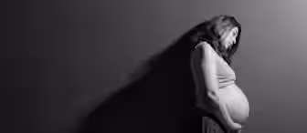 A black and white photo of a pregnant woman standing in profile against a plain wall. She is wearing a sleeveless shirt and pants, with one hand resting on her belly. The lighting creates a soft shadow on the wall behind her.