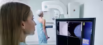 A healthcare professional reviews mammogram images on a computer monitor while a patient, standing and dressed in a hospital gown, undergoes a mammography examination in the background. The scene takes place in a medical imaging room.