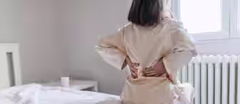 A person in light-colored pajamas sits on the edge of a bed, holding their lower back with both hands, indicating discomfort. The room is softly lit with a radiator and a cup on a nearby nightstand in the background.