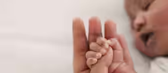 Close-up of a baby holding an adult's finger while resting on a white surface. The baby's small hand is grasping the adult's finger, showcasing the contrast in size and the bond between them. The baby's face is partially visible in the background, appearing to rest peacefully.