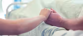A close-up image of two people holding hands, with one laying in a hospital bed covered by a light-colored blanket. The scene is gentle and supportive, indicating care and comfort, with the background softly blurred.