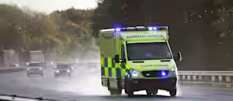 An ambulance with flashing lights drives quickly on a wet highway, passing other vehicles. Trees line the road and the sky is overcast, suggesting recent or ongoing rain.
