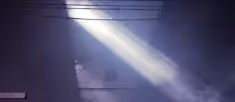 Light beams shine through the fog in an industrial setting, illuminating the metal framework and hanging light fixtures from a high ceiling. The scene evokes a sense of quiet and mystery in the dimly lit space.