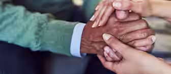 A close-up of two people's hands gently holding and comforting an older person's hands. The older person is wearing a green sweater, while the other individual’s hands are bare. The image conveys support, care, and compassion.
