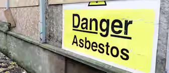 A yellow and white sign on a boarded-up building wall reads "Danger Asbestos," warning of hazardous materials inside.