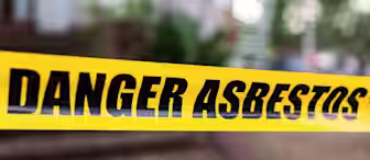 Yellow caution tape reading "Danger Asbestos" stretches across an area, warning of potential asbestos hazard. A blurred background shows an outdoor setting with trees and a building.