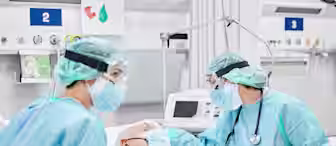 Two healthcare workers in full protective gear, including face shields, masks, gloves, and gowns, attend to an elderly patient lying in a hospital bed. Medical equipment and monitors are visible in the background. The environment appears to be an ICU.