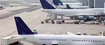Several large commercial airplanes are parked at the gates of a busy airport terminal. Various ground service vehicles are attending to the planes, and the airport runway can be seen in the background under a clear blue sky.