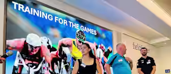 Four people with prosthetic limbs stand in front of a large screen displaying cyclists in a race, with the text "Training for the Games" above them. They appear to be in a conference room with a row of chairs.