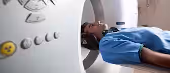 A person wearing a blue medical gown is lying on a table, entering an MRI or CT scanner. The scanner's control panel with various buttons is visible in the foreground. The room appears to be a medical facility with equipment on the walls.