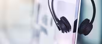A black headset with a microphone rests on the edge of a computer monitor. The background is softly blurred, suggesting a modern office environment. Natural light filters in, creating a bright and professional atmosphere.