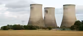 Three large, cylindrical cooling towers stand against a cloudy sky. They are surrounded by greenery and a field in the foreground. The towers are part of an industrial plant, possibly a power station.