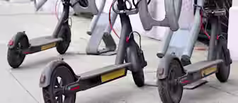 Three electric scooters parked on a gray tiled sidewalk. They are secured to metal stands with red cables. The scooters have black frames and sturdy wheels. The background features a light-colored wall.